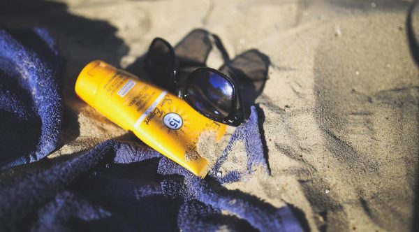 Eine Tube Sonnencreme liegt gemeinsam mit einer Sonnenbrille auf einem Handtuch im Sand.